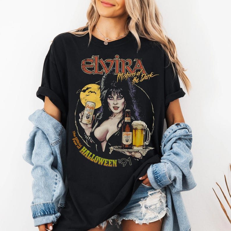 Op de afbeelding: Zwart T-shirt met een afbeelding van Elvira, Mistress of the Dark, met een biertje en een blikje bier. Het ontwerp bevat de tekst "The Official Fear of Halloween '91". Het ontwerp is in tinten rood, geel en wit.