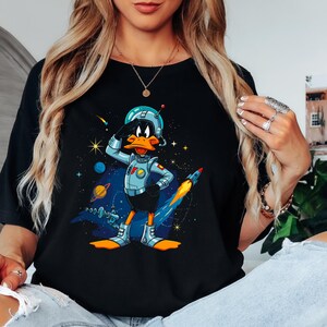 May include: Black t-shirt featuring a cartoon Daffy Duck in a space suit, saluting. The design includes planets, stars, and a rocket ship against a starry background. The duck is wearing a blue helmet and space suit.