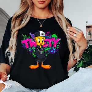 May include: Black t-shirt featuring a cartoon Tweety Bird character wearing a backwards cap and a hoodie with a silver chain. The word "TWEETY" is in pink and green graffiti-style lettering, with musical notes in the background.