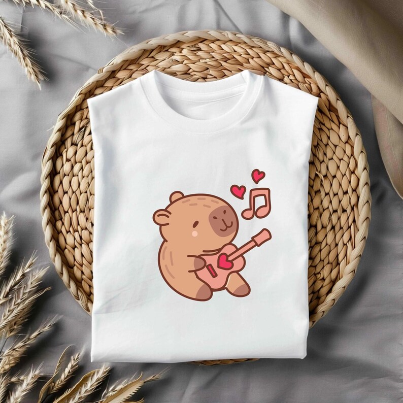 May include: White t-shirt with a cartoon bear playing a pink guitar. Two red hearts and musical notes are above the bear. The shirt is displayed on a woven basket. The guitar has a heart-shaped sound hole.
