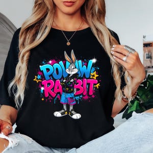 May include: Black t-shirt featuring a cartoon Bugs Bunny character with the words "POW RABBIT" in graffiti-style lettering. The graphic includes vibrant pink, blue, and yellow colors. Bugs Bunny is wearing a jacket, shorts, and sneakers.