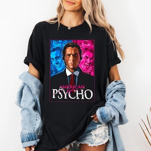 Op de afbeelding: Zwart t-shirt met een afbeelding uit de film "American Psycho". Het ontwerp bevat een portret van de hoofdpersoon met blauwe en roze accenten, en de titel van de film in witte letters.