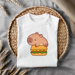 May include: White t-shirt with a cartoon capybara on a burger. The capybara is brown, and the burger has yellow, green, and brown layers. The shirt is displayed on a woven tray. A fun, casual design.