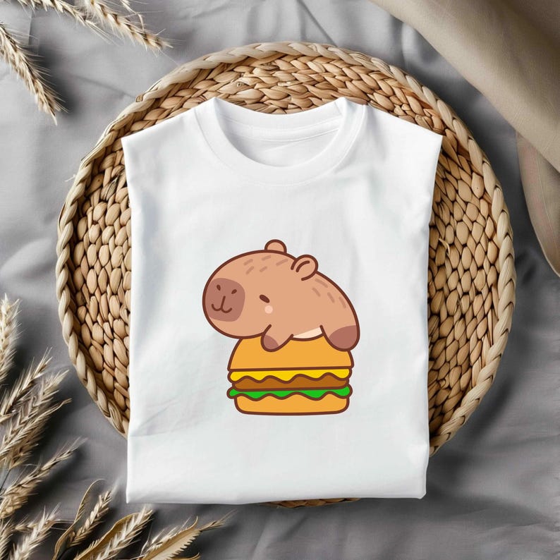 May include: White t-shirt with a cartoon capybara on a burger. The capybara is brown, and the burger has yellow, green, and brown layers. The shirt is displayed on a woven tray. A fun, casual design.
