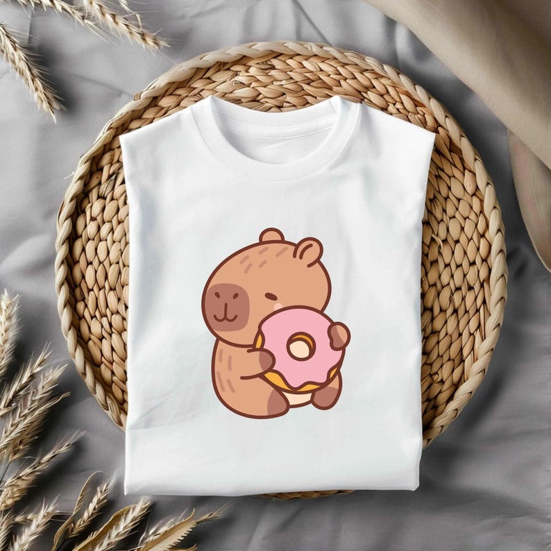 May include: White t-shirt with a cartoon capybara holding a pink donut. The capybara is brown and smiling. The donut has pink icing and a yellow centre. The t-shirt is folded on a woven tray, with a neutral background.