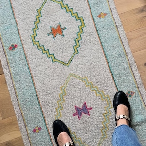 Könnte beinhalten: Ein rechteckiger, hellgrauer Teppich mit geometrischem Design. Der Teppich hat eine zentrale Diamantenform mit einem Sternmuster in Orange und Lila, umrandet von Gelb und Türkis. Der Rand ist türkis. Schwarze Schuhe mit dekorativen Riemen sind sichtbar.
