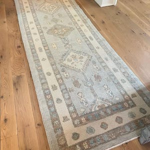 Könnte beinhalten: Ein rechteckiger, hellblauer und beigefarbener Läuferteppich mit einem komplizierten, sich wiederholenden Muster. Der Teppich hat ein zentrales Feld in Hellblau mit beige- und braunen Akzenten und einen Rand mit geometrischen Designs. Der Teppich liegt auf einem Holzboden.