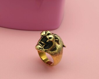 May include: A gold-colored ring featuring a roaring tiger head design. The ring has a detailed texture and a dark interior in the tiger's mouth. The ring is set against a pink background, with a pink container in the background.