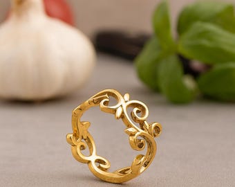 May include: A gold-colored ring with an ornate, swirling floral design. The ring features a series of interconnected curves and leaf-like shapes, creating a delicate and elegant aesthetic. The ring is set against a soft gray background.