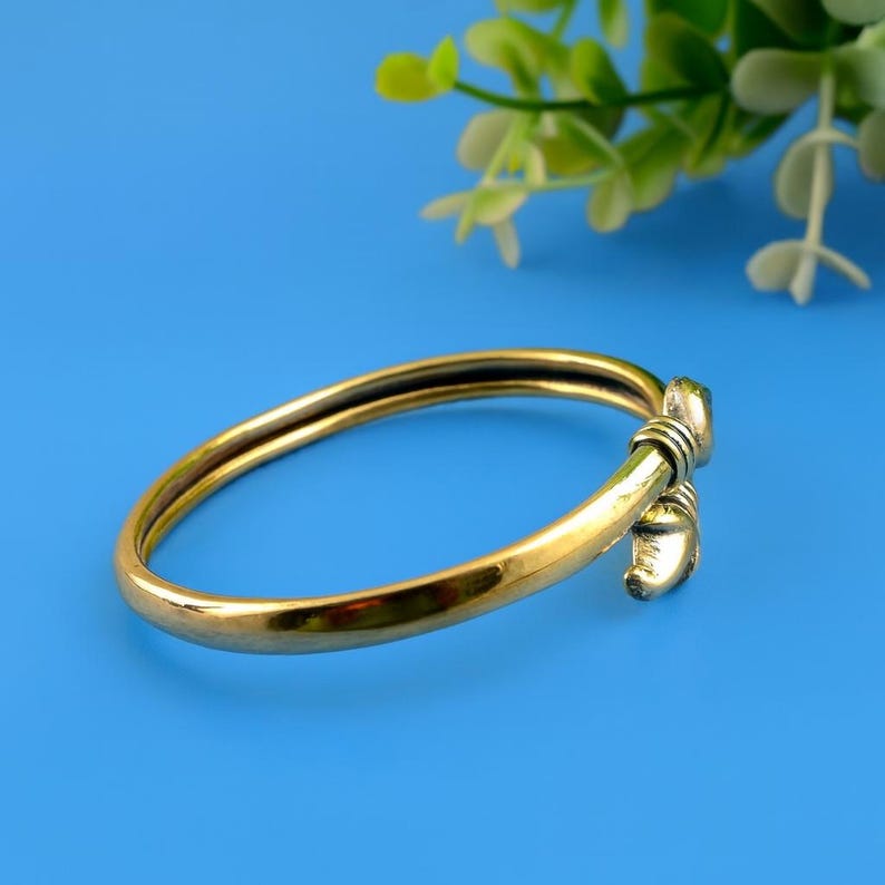 Puede incluir: Un brazalete de color dorado con un cierre decorativo. El brazalete tiene un acabado liso y pulido y un dise&ntilde;o simple y elegante. El cierre presenta un dise&ntilde;o detallado y texturizado. El fondo es de color azul liso.