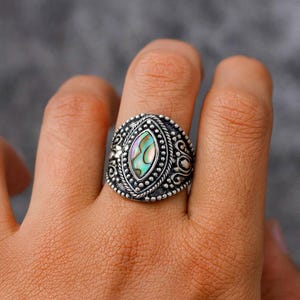 May include: A silver-toned ring with an oval, iridescent abalone shell center stone. The ring features intricate detailing, including a beaded border and decorative flourishes. The ring is a piece of jewelry.