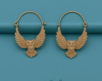 May include: A pair of gold-colored hoop earrings. Each earring features a detailed owl pendant with outstretched wings. The owls are facing forward, and the earrings are set against a teal background.