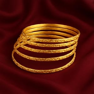 May include: A stack of six gold bangles with intricate floral and geometric patterns. The bangles are displayed on a deep red fabric, showcasing their bright, polished finish. Each bangle is approximately 0.25 inches wide.