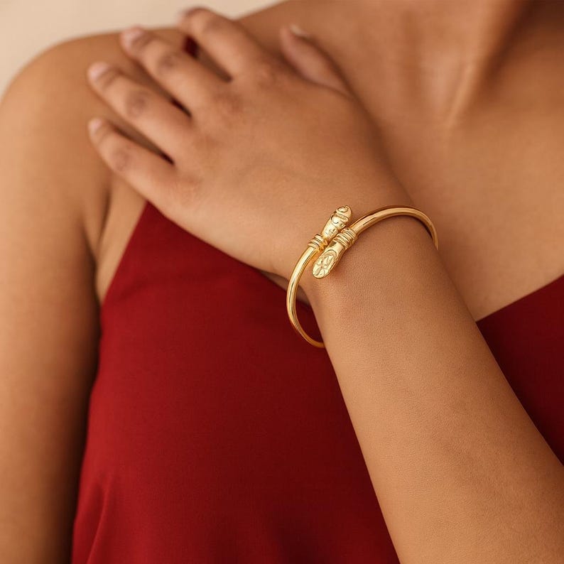 Puede incluir: Un brazalete dorado con extremos decorativos. El brazalete se lleva en la mu&ntilde;eca y los extremos presentan dise&ntilde;os detallados. El fondo es de color rojo liso.