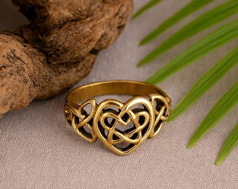 May include: A gold-coloured ring with a Celtic knot heart design. The ring features an intricate, interwoven pattern forming a heart shape in the centre. The ring is displayed on a neutral surface with a wooden element and green leaves in the background.