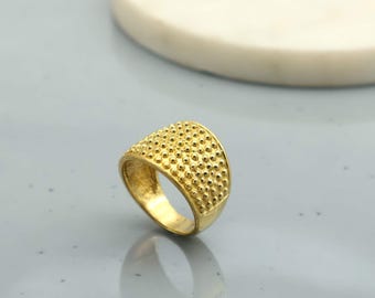 May include: A gold-colored ring with a textured surface of small, evenly spaced bumps. The ring has a wide band and a rounded top, reflecting light. The ring is likely made of metal and is displayed on a light gray surface.