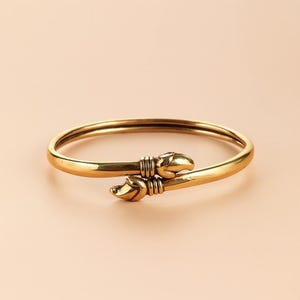 Puede incluir: Una pulsera dorada con un dise&ntilde;o &uacute;nico. La pulsera presenta una banda lisa y redondeada que se estrecha hacia dos extremos decorativos. Cada extremo tiene forma de garra, lo que a&ntilde;ade un toque distintivo a la joya. La pulsera est&aacute; sobre un fondo neutro.