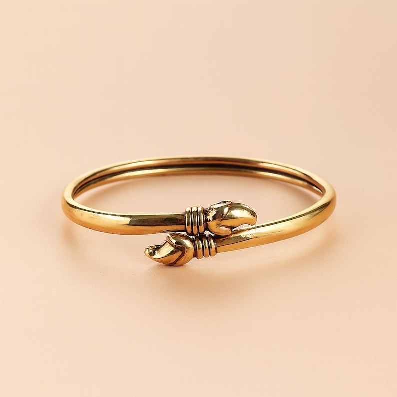 Puede incluir: Una pulsera dorada con un dise&ntilde;o &uacute;nico. La pulsera presenta una banda lisa y redondeada que se estrecha hacia dos extremos decorativos. Cada extremo tiene forma de garra, lo que a&ntilde;ade un toque distintivo a la joya. La pulsera est&aacute; sobre un fondo neutro.