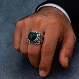 Könnte beinhalten: Ein silberner Ring mit einem großen, runden, dunkelgrünen Stein. Der Ring hat ein verziertes, detailliertes Design um den Stein herum. Der Ring befindet sich an einem Finger.