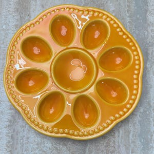 May include: A ceramic egg serving platter in a warm orange hue. The platter features eight egg-shaped indentations around a central circular well. The rim is adorned with a beaded design, adding a touch of elegance to this serving dish.