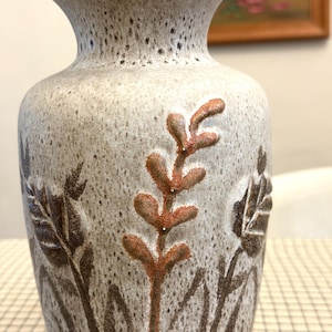 Puede incluir: Un jarrón de cerámica con un acabado gris claro moteado. El jarrón presenta diseños botánicos marrones y marrón rojizo en relieve. El jarrón tiene una boca ancha y una superficie texturizada, adecuado para exhibir flores o como pieza decorativa.