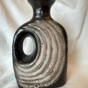 Könnte beinhalten: Dunkelbraune Keramikvase mit einzigartigem Design. Die Vase hat eine kreisförmige Öffnung und ein strukturiertes, geschichtetes Muster in Weiß und Braun. Die Vase hat einen schmalen Hals und einen abgerundeten Körper, ideal für die Inneneinrichtung. Ca. 25 cm hoch.