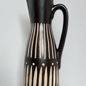 Puede incluir: Una jarra de cerámica marrón oscuro con asa y boquilla. La jarra está decorada con rayas verticales de color crema y pequeños puntos. Esta jarra decorativa es adecuada para servir agua u otras bebidas.