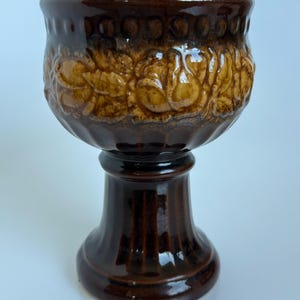 Può includere: Un vaso a piedistallo in ceramica marrone con finitura lucida. La parte superiore presenta un motivo floreale in rilievo strutturato in una tonalità marrone più chiara. Il vaso ha una parte superiore ampia e arrotondata e una base scanalata.