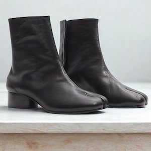 Op de afbeelding: Een paar zwarte leren enkellaarsjes met een split-teenontwerp. De laarzen hebben een korte blokhak en een ritssluiting aan de zijkant. De laarzen worden op een wit oppervlak weergegeven.