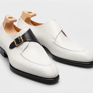 May include: A pair of white leather monk strap shoes featuring a contrasting black leather strap with a gold-toned metal buckle, a black sole, and light-colored wooden shoe trees inserted into the heel area of each shoe.