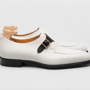 May include: A white leather single monk strap shoe featuring a black leather strap with a gold-toned metal buckle, a black sole, and a light-colored wooden shoe tree inserted into the heel area to maintain the shape of the footwear.