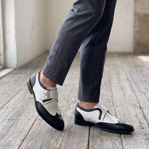 May include: Two-tone leather shoes with a black toe and heel, and white upper. The shoes feature a buckle closure and are worn with gray trousers. The shoes are on a wooden floor.