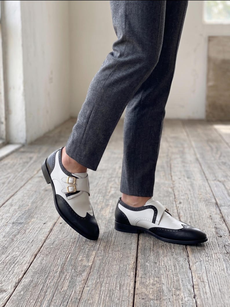 May include: Two-tone leather shoes with a black toe and heel, and white upper. The shoes feature a buckle closure and are worn with gray trousers. The shoes are on a wooden floor.