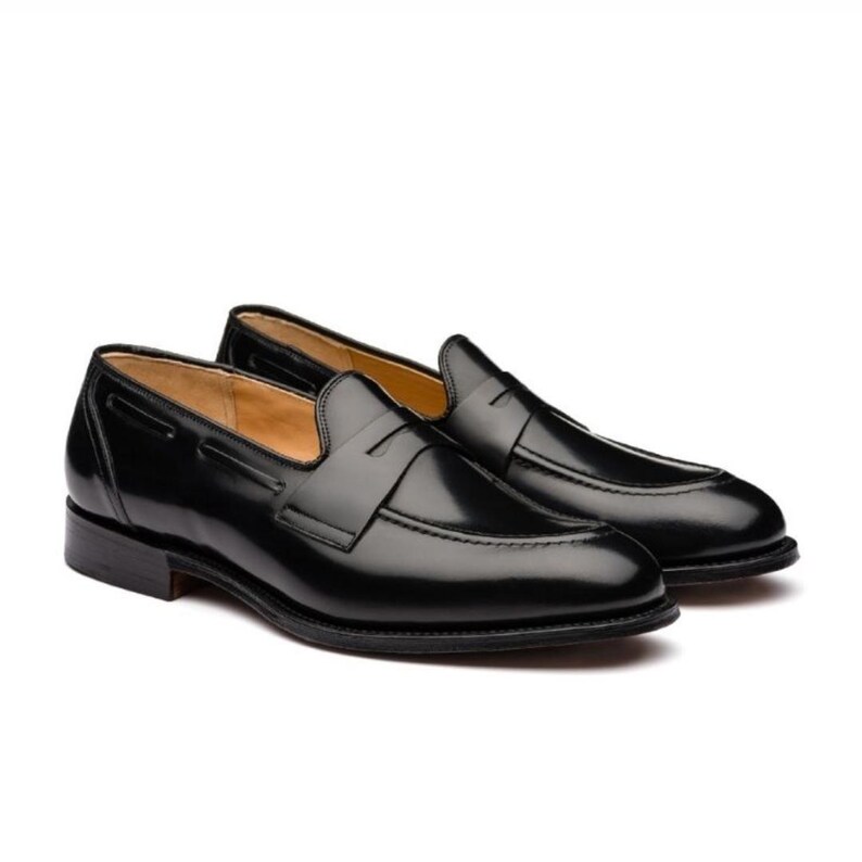 May include: A pair of black leather penny loafers. The shoes have a glossy finish and a classic design, featuring a strap across the vamp. The soles are dark, and the interior is a lighter shade. The shoes are set against a white background.