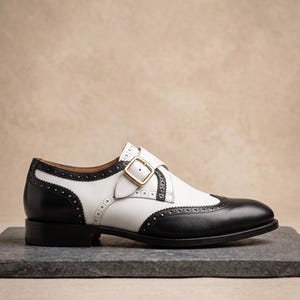 May include: A two-tone leather shoe with a black toe and heel, and white midsection. The shoe features a buckle closure with gold hardware and decorative perforations along the edges. The shoe is set on a dark gray surface.