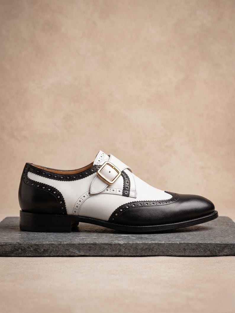 May include: A two-tone leather shoe with a black toe and heel, and white midsection. The shoe features a buckle closure with gold hardware and decorative perforations along the edges. The shoe is set on a dark gray surface.