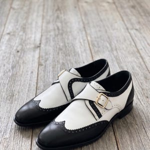 May include: A pair of two-tone leather shoes with black and white panels. The shoes feature a decorative perforated design along the edges and a gold-colored buckle closure. The shoes are displayed on a wooden surface.