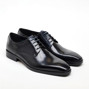 Può includere: Un paio di scarpe eleganti in pelle nera con finitura lucida. Le scarpe hanno un design classico con tacco basso, punta arrotondata e lacci neri. I lacci sono annodati. Su sfondo bianco.