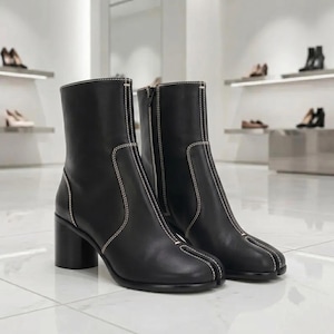 May include: A pair of black leather ankle boots featuring a distinct split-toe design, a block heel, and visible white contrast stitching along the seams. The boots have a side zipper closure and a smooth finish, presented in a clean, bright setting.