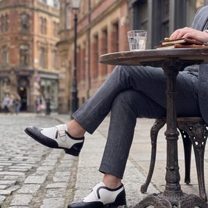 May include: Black and white leather shoes with a buckle closure. The shoes have a wingtip design and are worn with gray trousers. The setting is an outdoor cafe with a cobblestone street and historic buildings in the background.