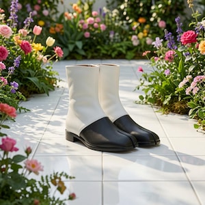 May include: A pair of ankle boots featuring a two-tone design with a white upper shaft and a black lower section. The boots have a distinct split-toe construction, a low block heel, and a side zipper closure, presenting a clean and structured look.