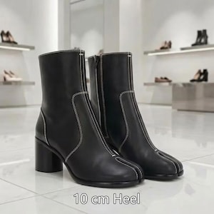 May include: A pair of black ankle boots featuring a split-toe design, white contrast stitching, a side zipper closure, and a block heel measuring 10 cm in height. The boots have a smooth finish and a structured silhouette with a distinct toe separation.
