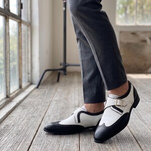 May include: Black and white leather dress shoes with a buckle closure. The shoes have a wingtip design with black accents and a white upper. The shoes are worn with gray trousers.