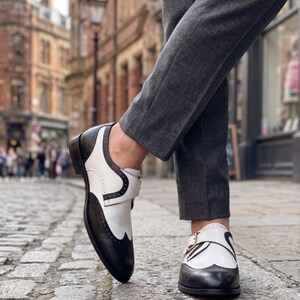 May include: Two-tone black and white leather shoes with a wingtip design. The shoes feature a buckle closure and are worn with gray trousers. The shoes are on a cobblestone street, with a blurred background of buildings.