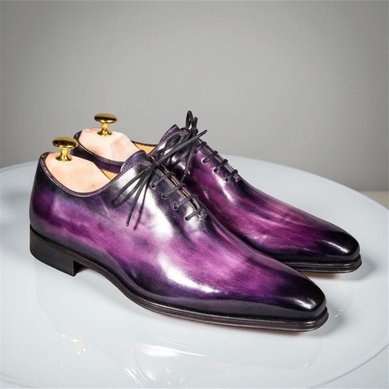 May include: A pair of purple leather dress shoes with black laces. The shoes have a glossy finish and a classic Oxford design. The toe of each shoe is a darker shade of purple. Shoe trees are inside the shoes.