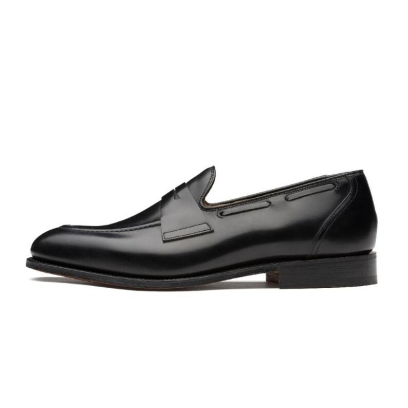 May include: A single black leather loafer shoe. The shoe has a sleek, polished appearance with a low heel and a decorative strap across the vamp. The design is classic and suitable for formal or business attire.