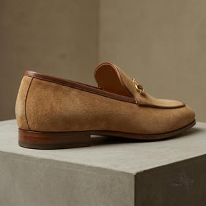 Pu&ograve; includere: Un mocassino in camoscio color cuoio con un bordo in pelle marrone liscia lungo il colletto e un dettaglio morsetto in metallo dorato sul davanti. La scarpa presenta un tacco basso in legno a strati e una punta arrotondata e classica.