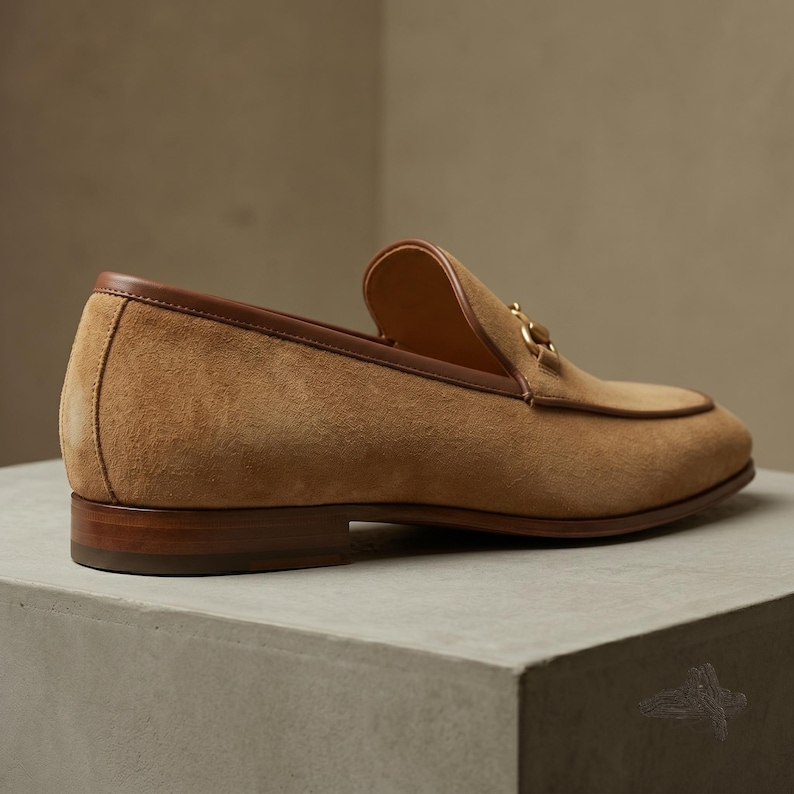 Pu&ograve; includere: Un mocassino in camoscio color cuoio con un bordo in pelle marrone liscia lungo il colletto e un dettaglio morsetto in metallo dorato sul davanti. La scarpa presenta un tacco basso in legno a strati e una punta arrotondata e classica.