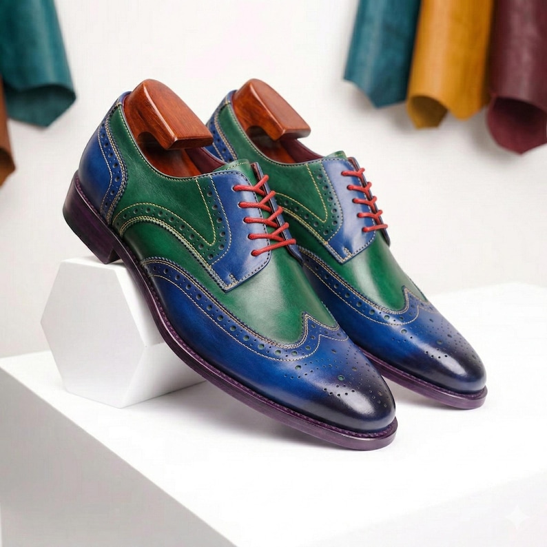 Op de afbeelding: Tweekleurige leren schoenen in blauw en groen met rode veters. De schoenen worden gepresenteerd op een witte ondergrond. De schoenen hebben houten schoenspanners.