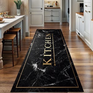 Può includere: Un tappeto da cucina rettangolare con un design in marmo nero e bordi dorati. La parola "KITCHEN" è visualizzata verticalmente in lettere dorate. Il tappeto è posizionato su un pavimento in legno.